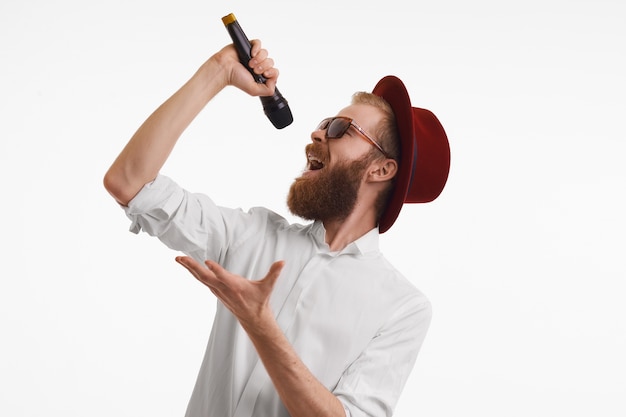 Persone, musica, divertimento, spettacolo e concetto di intrattenimento. artista pop dai capelli rossi elegante bello emotivo con folta barba che canta nel microfono che indossa un cappello rotondo rosso e sfumature alla moda