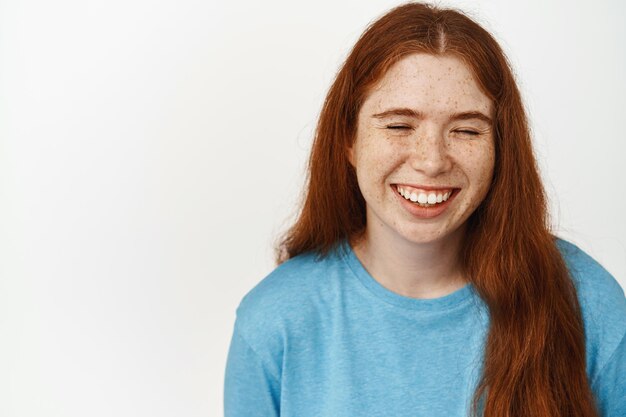 Persone positive. Giovane ragazza felice con i capelli lunghi rossi, che ride spensierata con gli occhi chiusi, in piedi in maglietta blu su sfondo bianco