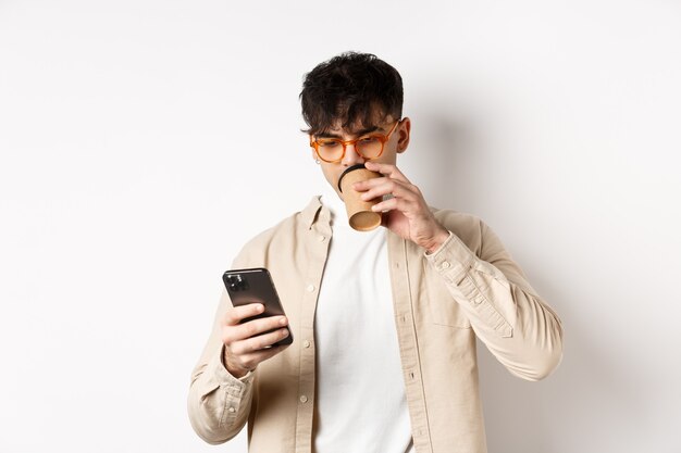 Persone reali. Giovane che legge lo schermo dello smartphone e beve caffè, guardando il telefono, in piedi su sfondo bianco