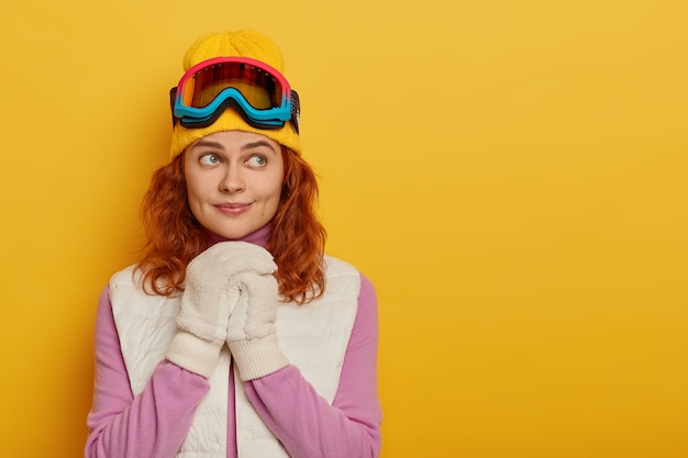 Persone, ricreazione, pensieri, concetto di attività per il tempo libero. Adorabile donna allo zenzero tiene le mani unite sul petto, indossa abiti caldi, maschera da snowboard, pensa a una nuova avventura durante l'inverno