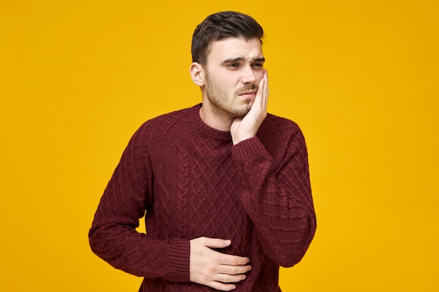 Persone, sanità, odontoiatria e concetto di malattia. Giovane uomo con la barba lunga sconvolto depresso con espressione facciale dolorosa stressata che soffre di mal di denti sensazione di malessere, tenendo la mano sulla guancia e sullo stomaco