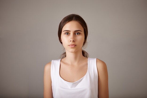 Persone, stile di vita, bellezza e concetto di moda. Chiuda sul ritratto di bella giovane posa femminile europea alla moda isolata avendo espressione facciale pacifica calma che porta la parte superiore senza maniche bianca