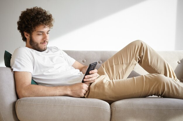 Persone, stile di vita moderno e concetto di gadget. Ragazzo alla moda hipster con barba sfocata e acconciatura riccia rilassante al chiuso, sdraiato sul divano con smart phone, godendo della comunicazione online, utilizzando il wifi