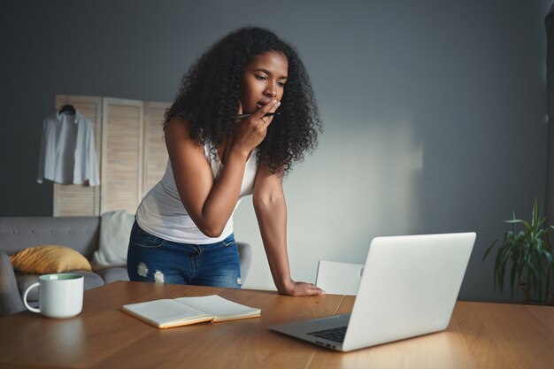 Persone, stile di vita, tecnologia moderna, concetto di lavoro e occupazione. Ritratto di una bellissima giovane scrittrice africana che si sente preoccupata, sperimentando il blocco creativo, utilizzando il laptop e prendendo appunti