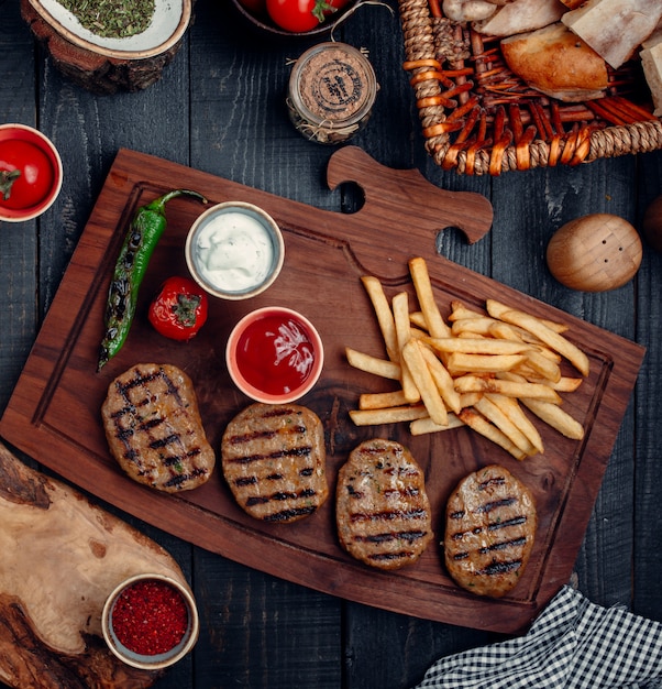 Pezzi di bistecca con patatine fritte, pepe e pomodoro grigliati e salse su una tavola di legno.