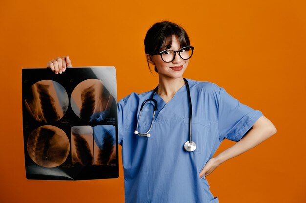 Piacere di mettere la mano sull'anca che tiene xray giovane dottoressa che indossa uno stetoscopio fith uniforme isolato su sfondo arancione