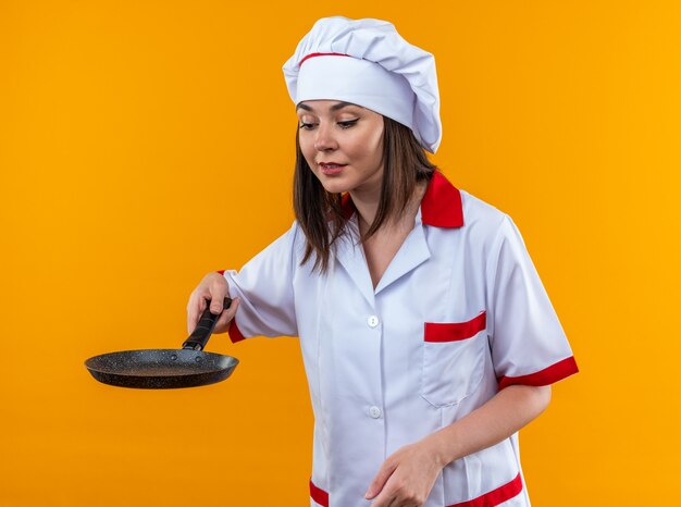 Piacere giovane cuoca che indossa l'uniforme dello chef tenendo e guardando la padella isolata su sfondo arancione