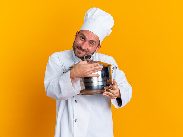 Piacere giovane maschio caucasico cuoco in uniforme da chef e cappello che tiene e abbraccia pentola