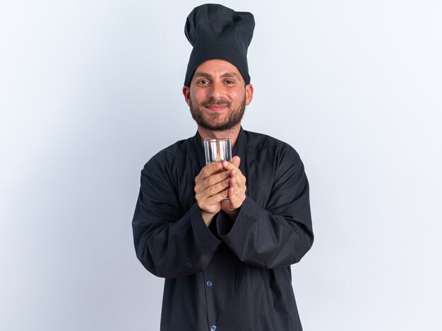 Piacere giovane maschio caucasico cuoco in uniforme da chef e cappuccio tenendo un bicchiere d'acqua guardando la telecamera isolata sul muro bianco con spazio di copia