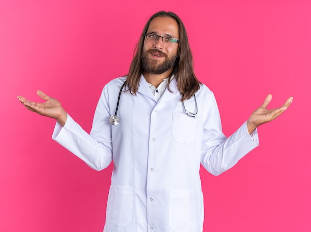 Piacere maschio adulto medico indossando accappatoio medico e stetoscopio con gli occhiali guardando la telecamera che mostra le mani vuote isolate sulla parete rosa