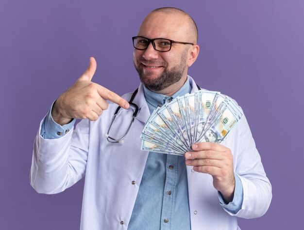 Piacere medico maschio di mezza età che indossa una tunica medica e uno stetoscopio con occhiali che tengono e puntano al denaro