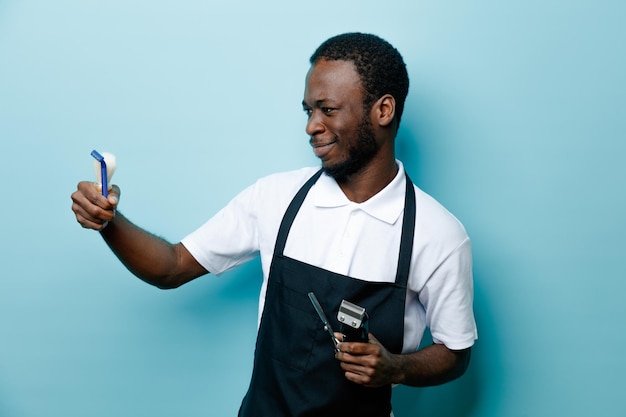 Piacevole azienda barbiere strumenti giovane barbiere afroamericano in uniforme isolato su sfondo blu