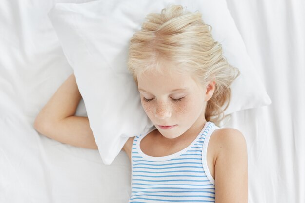 Piacevole bambina con i capelli biondi e la faccia lentigginosa, che dorme dolcemente sdraiata sul cuscino bianco chiudendo gli occhi, godendo di un'atmosfera calma e di condizioni confortevoli nella sua camera da letto