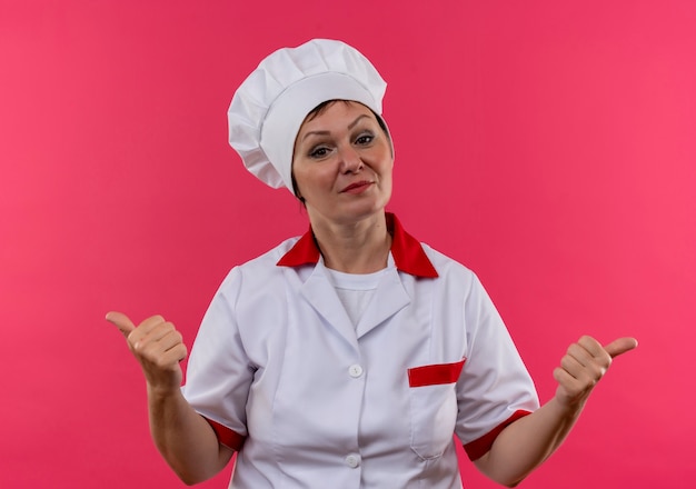 Piacevole cuoca di mezza età in uniforme da chef i suoi pollici in su sulla parete rosa isolata