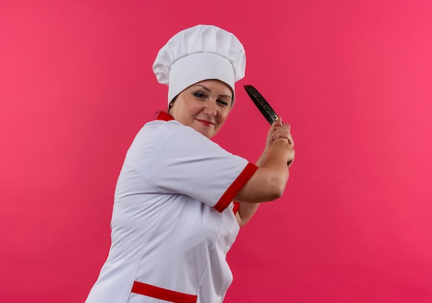 Piacevole cuoca di mezza età in uniforme del cuoco unico che tiene la padella intorno alla spalla sulla parete rosa isolata con lo spazio della copia