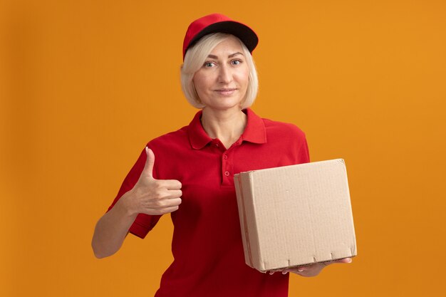 Piacevole donna bionda di mezza età in uniforme rossa e cappuccio che tiene in mano una scatola di cartone guardando la parte anteriore che mostra il pollice in alto isolato sulla parete arancione