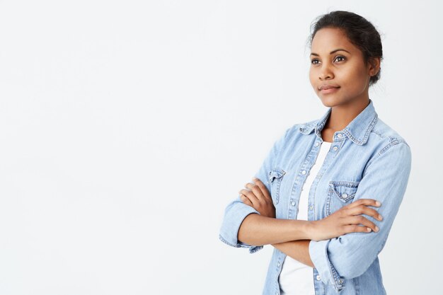 Piacevole donna dalla pelle scura con blackhair, occhi scuri vestiti con maglietta bianca, giacca di jeans che tengono le mani piegate sorridendo leggermente mentre posa sul muro bianco. Persone e stile di vita