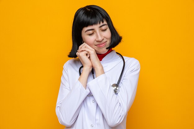 Piacevole giovane bella ragazza caucasica in uniforme da medico con stetoscopio che tiene le mani insieme in piedi con gli occhi chiusi