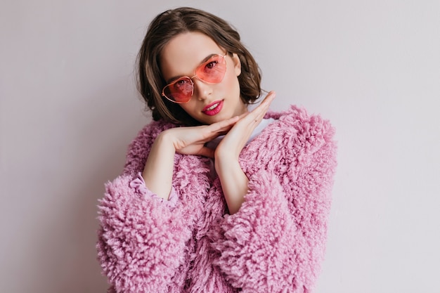 Piacevole giovane donna dai capelli castani in occhiali da sole luminosi chillng durante il servizio fotografico. Ritratto dell'interno di bella ragazza caucasica in cappotto di pelliccia che propone con interesse.