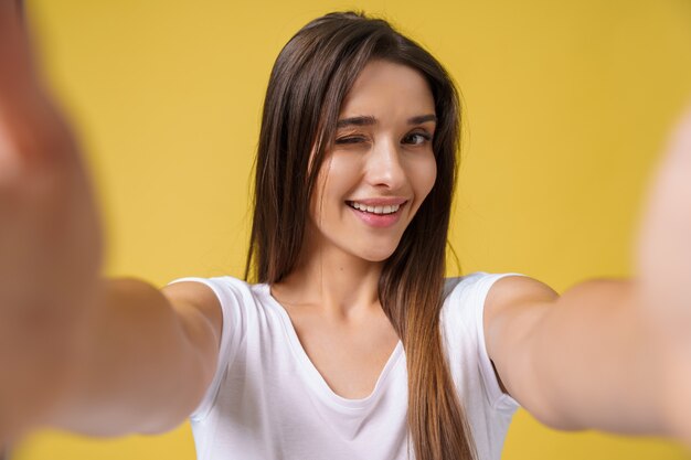 Piacevole ragazza attraente che fa selfie in studio e ridendo. Bella giovane donna con capelli castani che si fotografa su sfondo giallo brillante.