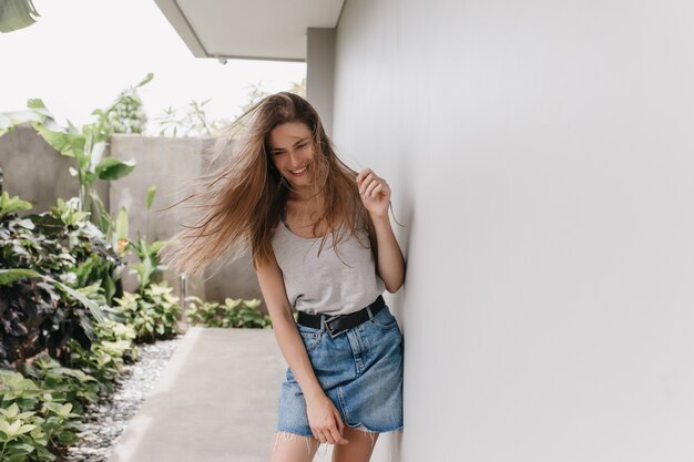 Piacevole ragazza in posa con i capelli lunghi agitando e ridendo accanto al muro bianco. Allegro modello femminile in gonna di jeans trascorrere del tempo all'aperto in una giornata calda.