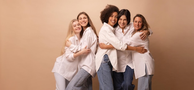 Piacevoli cinque diverse donne giovani e adulte in abbigliamento casual che si abbracciano sorridendo alla telecamera su sfondo marrone