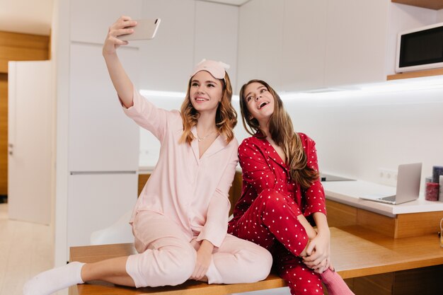 Piacevoli ragazze europee che fanno selfie prima di colazione. Tiro al coperto di bella bionda giovane donna di scattare una foto con il telefono in cucina.