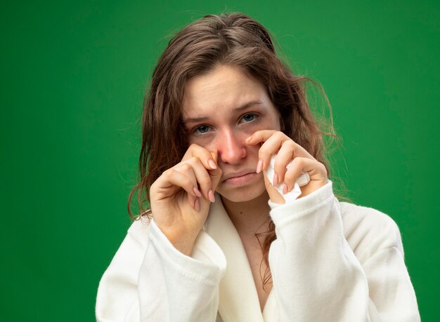 Piangere giovane ragazza malata che indossa una veste bianca asciugandosi gli occhi con le mani isolate su verde