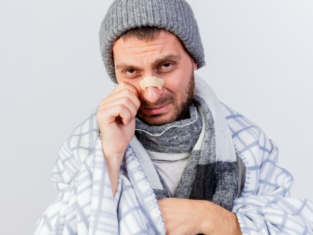 Piangere giovane uomo malato che indossa cappello invernale e sciarpa avvolto in un plaid con intonaco sul naso isolato su sfondo bianco