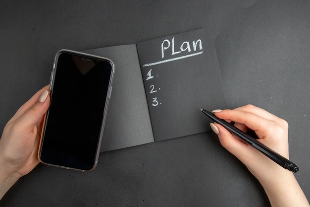 Piano vista dall'alto scritto sul telefono con blocco note nero e penna in mani femminili su sfondo nero