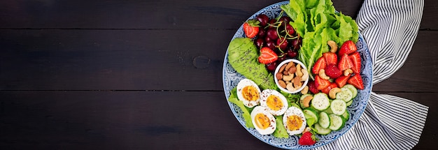 Piastra con un alimento dieta paleo. Uova sode, avocado, cetriolo, noci, ciliegia e fragole. Colazione Paleo. Vista dall'alto