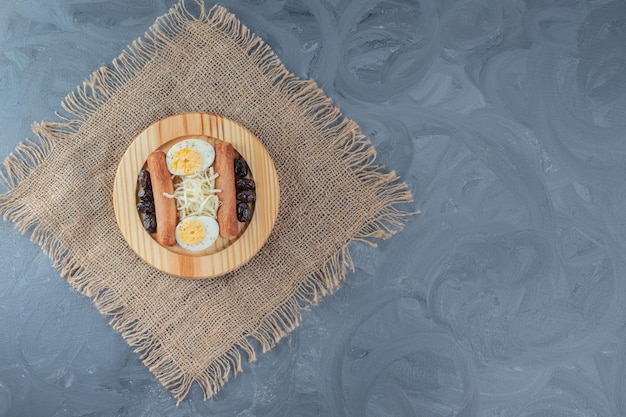 Piatto di legno con salsicce, uova a fette, formaggio grattugiato e olive nere su un pezzo di tessuto sul tavolo di marmo.