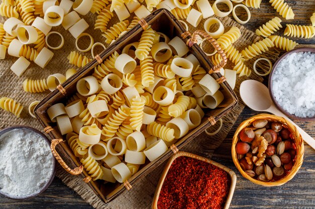 Piatto maccheroni pasta maccheroni nel vassoio con cucchiai, sale, spezie rosse, varie noci su tela di sacco e fondo in legno. orizzontale