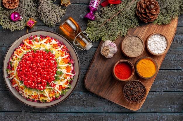 Piatto vista dall'alto e piatto di spezie con melograni accanto alla bottiglia di olio di spezie aglio sul tagliere e rami di albero con coni e giocattoli dell'albero di Natale