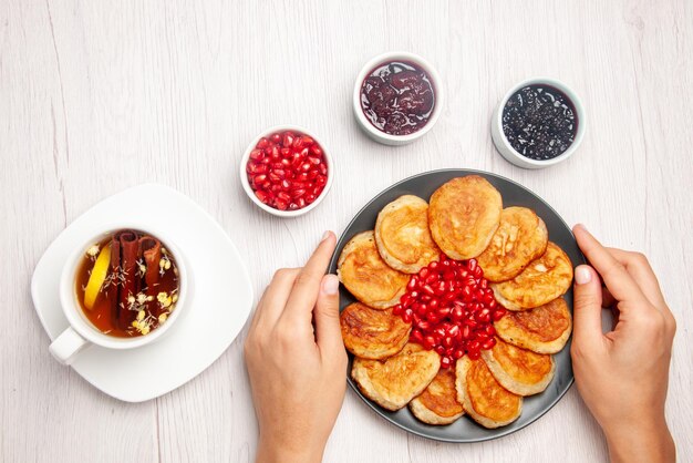 Piatto vista dall'alto in mano tazza di tè bianca con bastoncini di cannella ciotole di marmellata e piatto di frittelle e melograno in mano sul tavolo