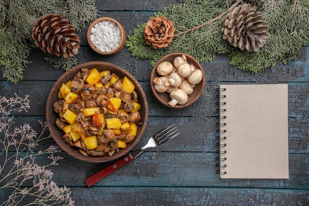 Piatto vista ravvicinata dall'alto e piatto di rami di funghi e patate sul tavolo grigio sotto i rami di abete con coni funghi e sale accanto alla forchetta e al taccuino