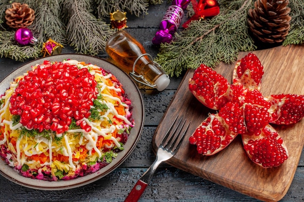 Piatto vista ravvicinata laterale e piatto di spezie con patate al melograno accanto alla bottiglia di olio di melograno forchetta sul tagliere e rami di albero con coni e giocattoli dell'albero di Natale