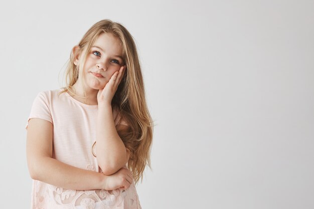 Piccola bella ragazza bionda con gli occhi azzurri che schiaccia il viso con la mano, guardando da parte con espressione frustrata dopo che la mamma conosce cattivi voti a scuola.