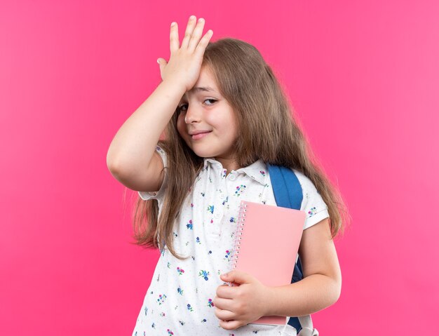 Piccola bella ragazza con i capelli lunghi con lo zaino che tiene il taccuino che sembra confusa tenendosi la mano sulla fronte per errore in piedi sul rosa