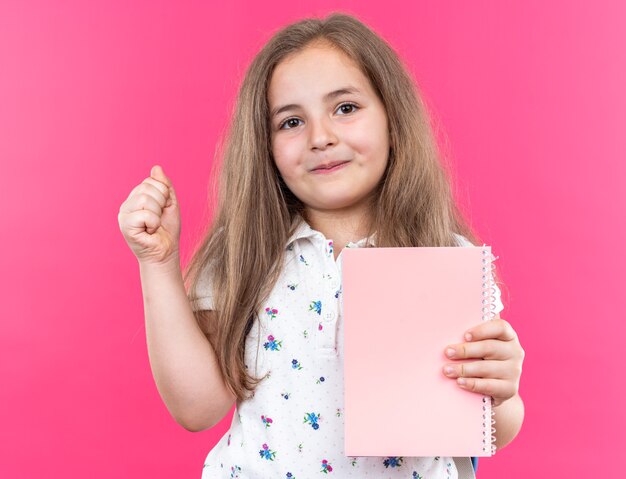 Piccola bella ragazza con i capelli lunghi con lo zaino che tiene il taccuino guardando davanti con un sorriso sul viso felice che mostra il pollice in piedi sul muro rosa