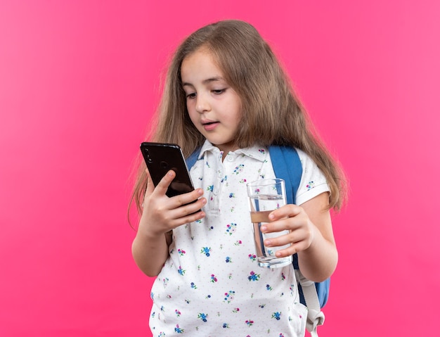 Piccola bella ragazza con i capelli lunghi con lo zaino che tiene smartphone e bicchiere d'acqua sorridente fiduciosa in piedi sul muro rosa