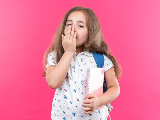 Piccola bella ragazza con i capelli lunghi con lo zaino che tiene smartphone guardando davanti stanco e annoiato che sbadiglia coprendo la bocca con la mano in piedi sul muro rosa