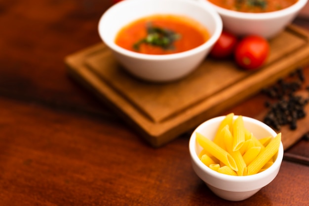 Piccola ciotola di pasta cruda del penne sulla tavola con le salse e i pomodori defocused sul bordo di legno