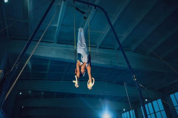 Piccola ginnasta maschio che si allena in palestra, flessibile e attiva. Ragazzo in forma caucasica, atleta in abiti sportivi bianchi che praticano esercizi per l'equilibrio sugli anelli.