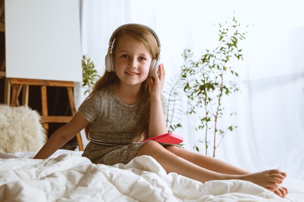 Piccola modella seduta nel suo letto con grandi cuffie ascoltando la musica preferita e godendo.