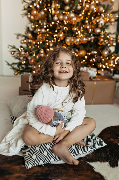 Piccola ragazza affascinante che si siede sul cuscino con il giocattolo sopra l'albero di Natale, umore di Capodanno, festa di Natale