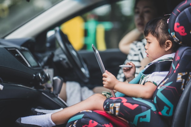 Piccola ragazza asiatica sveglia che si siede in una macchina con la sua mamma e che guarda i cartoni animati