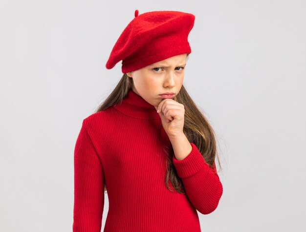 Piccola ragazza bionda accigliata che indossa un berretto rosso che tiene la mano sul mento isolato sul muro bianco con spazio per le copie