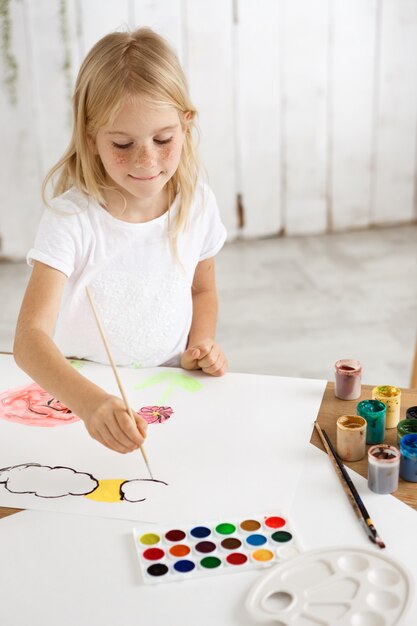 Piccola ragazza bionda allegra e adorabile con le lentiggini in panno bianco che disegna le nuvole e i fiori sul foglio di carta bianco