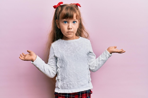 Piccola ragazza caucasica che indossa abiti casual all'oscuro e confusa con le braccia aperte, nessuna idea e faccia dubbiosa.